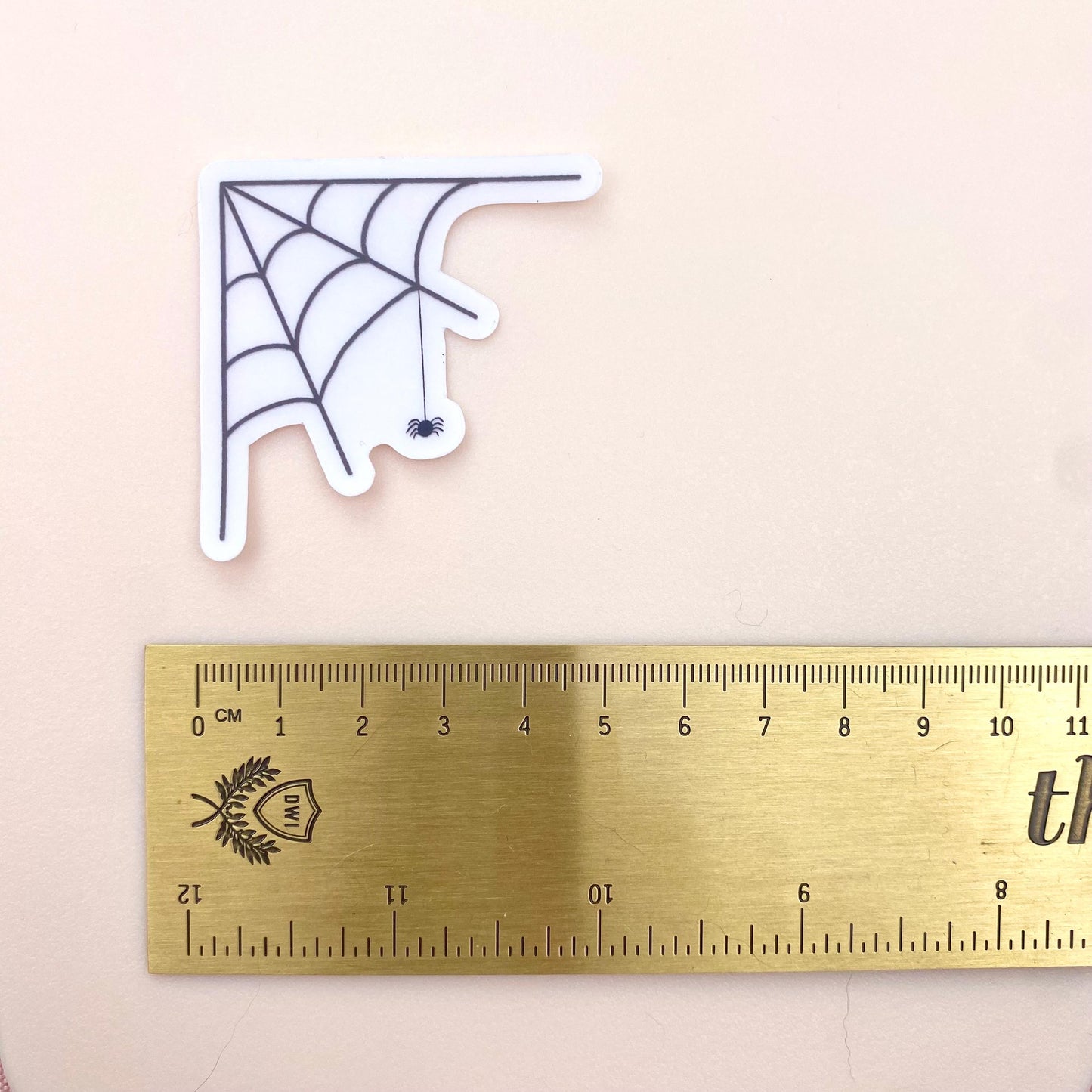 Transparent sticker with corner spider web design with a hanging spider on a white background with a gold ruler for scale.