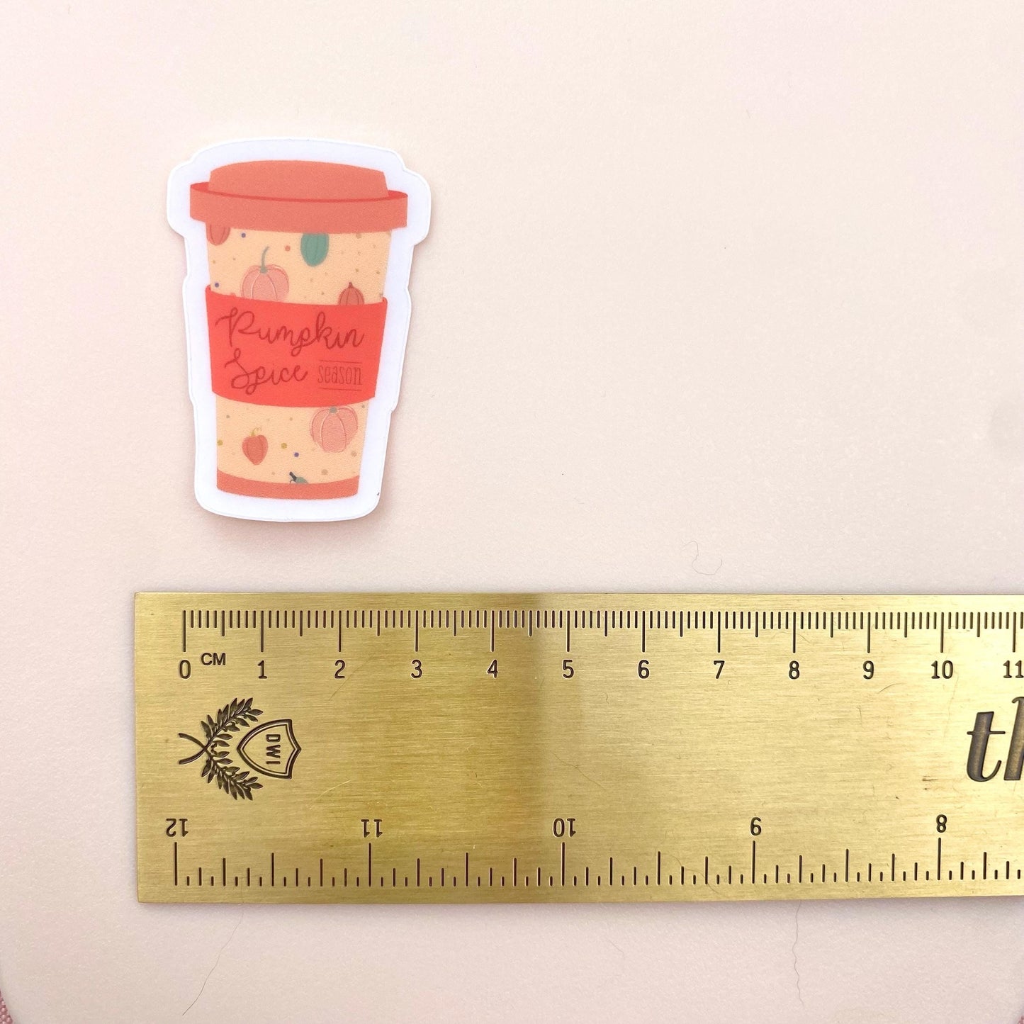 Sticker of a pumpkin spice latte cup next to a ruler on a beige background