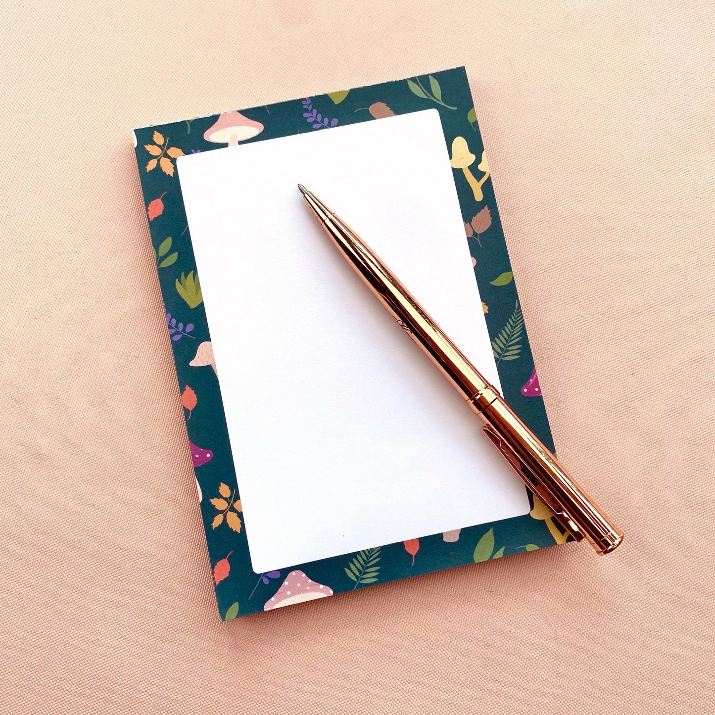 Notepad with mushroom and leaf patterned border on a white background with a rose gold pen