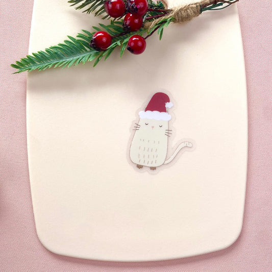Decorative cat sticker with Santa hat on a white board with holly leaves and berries.