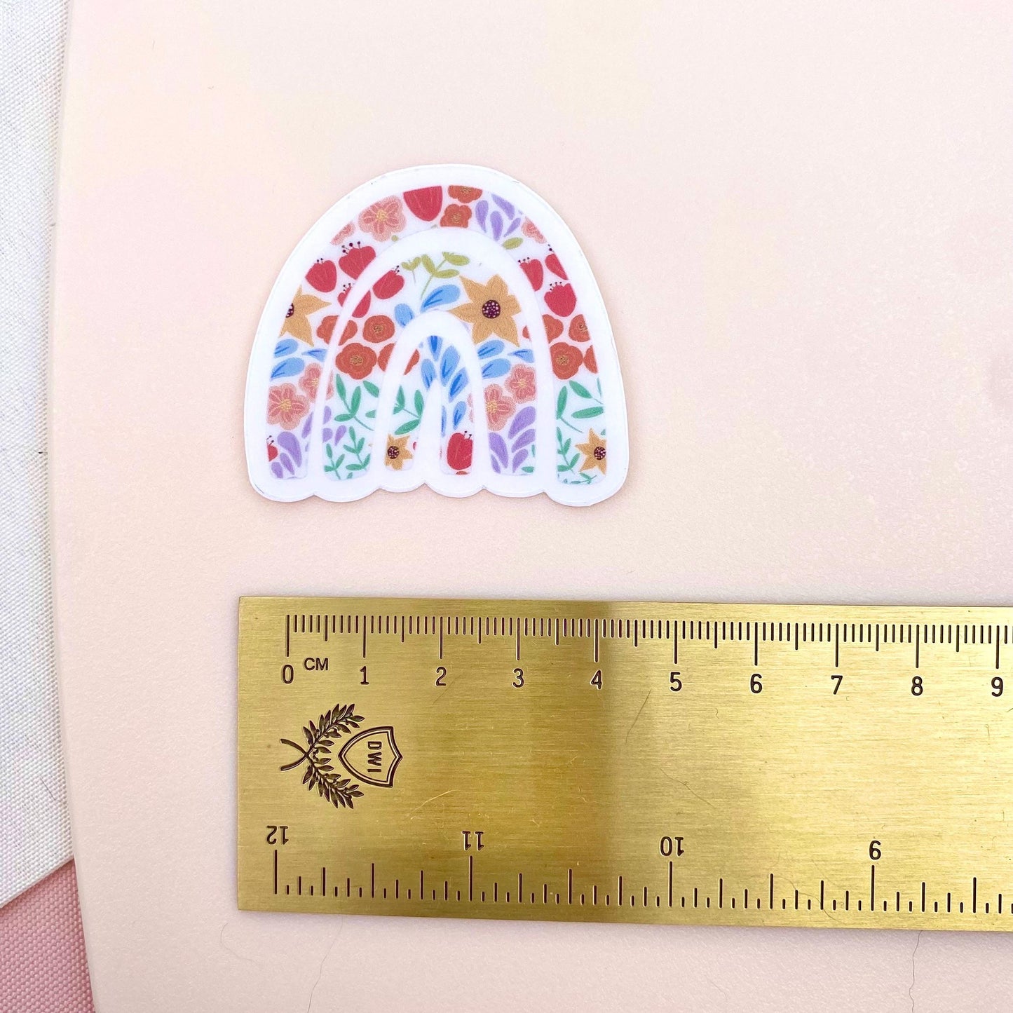 Decorative rainbow sticker with floral design next to a ruler on a light pink background