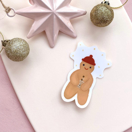 Sticker of a gingerbread man with a red woolly hat on a pink background with Christmas decorations.