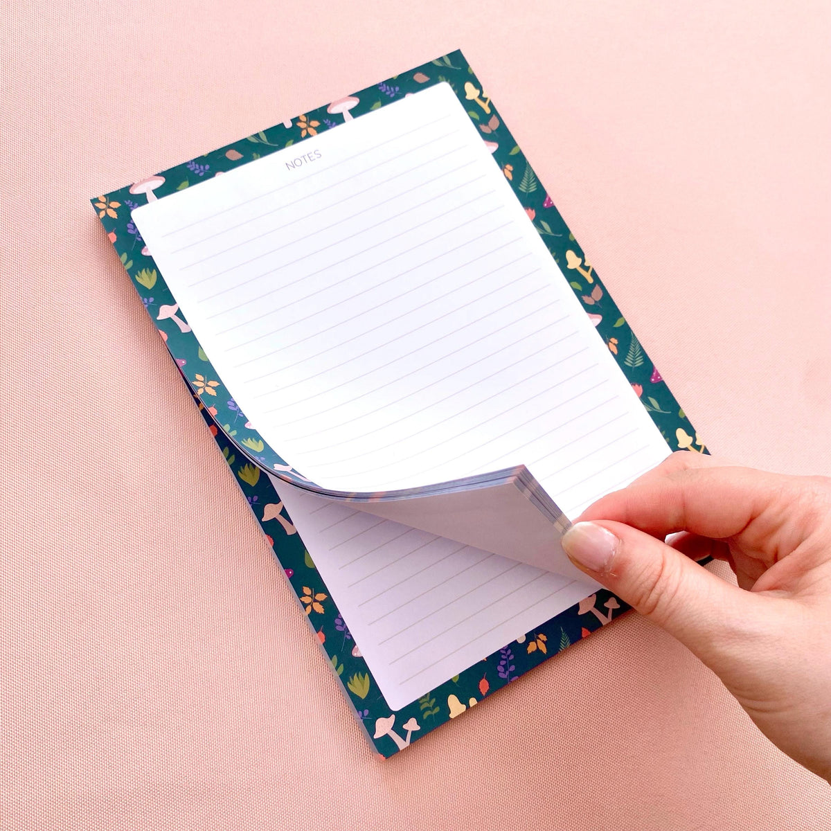 Notepad with autumn foliage border held open by a hand on a pink background