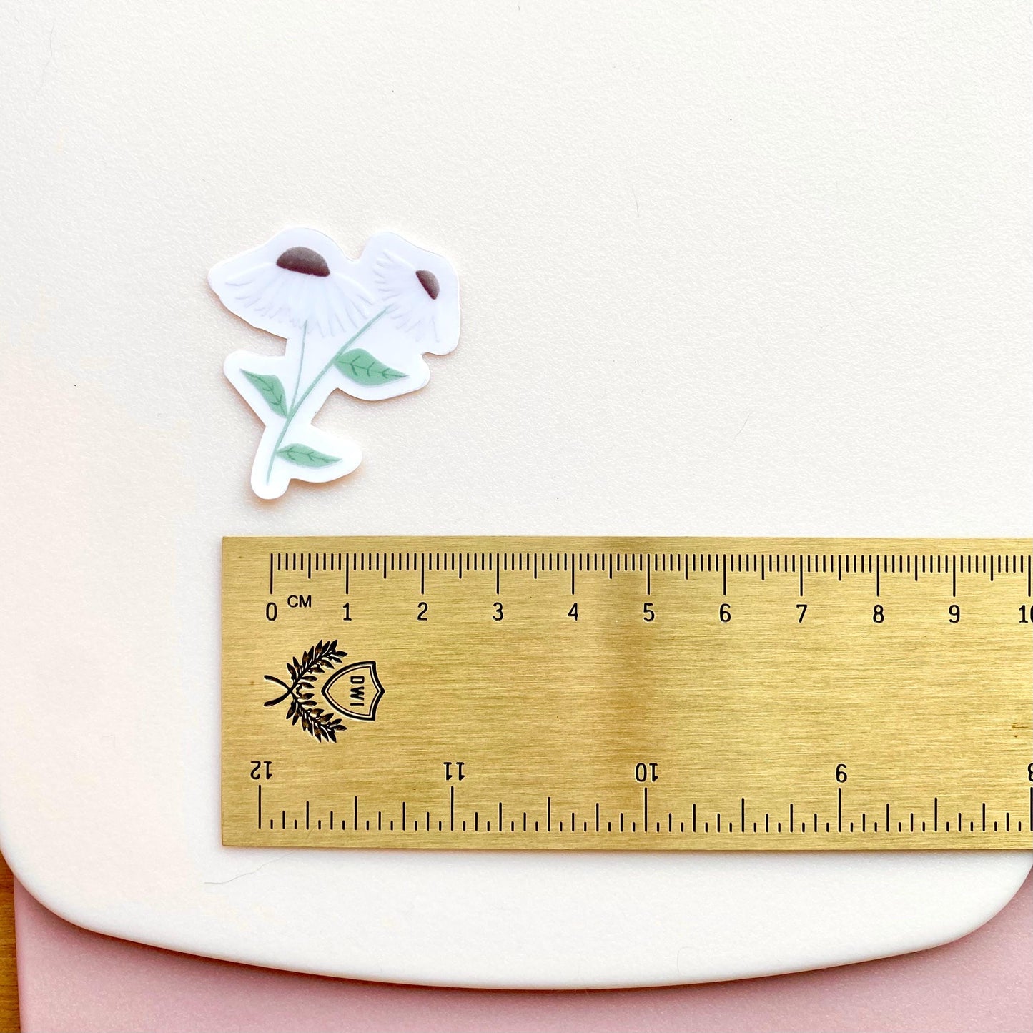 A clear sticker with daisies with white petals and green leaves on a white background with a gold ruler for scale.