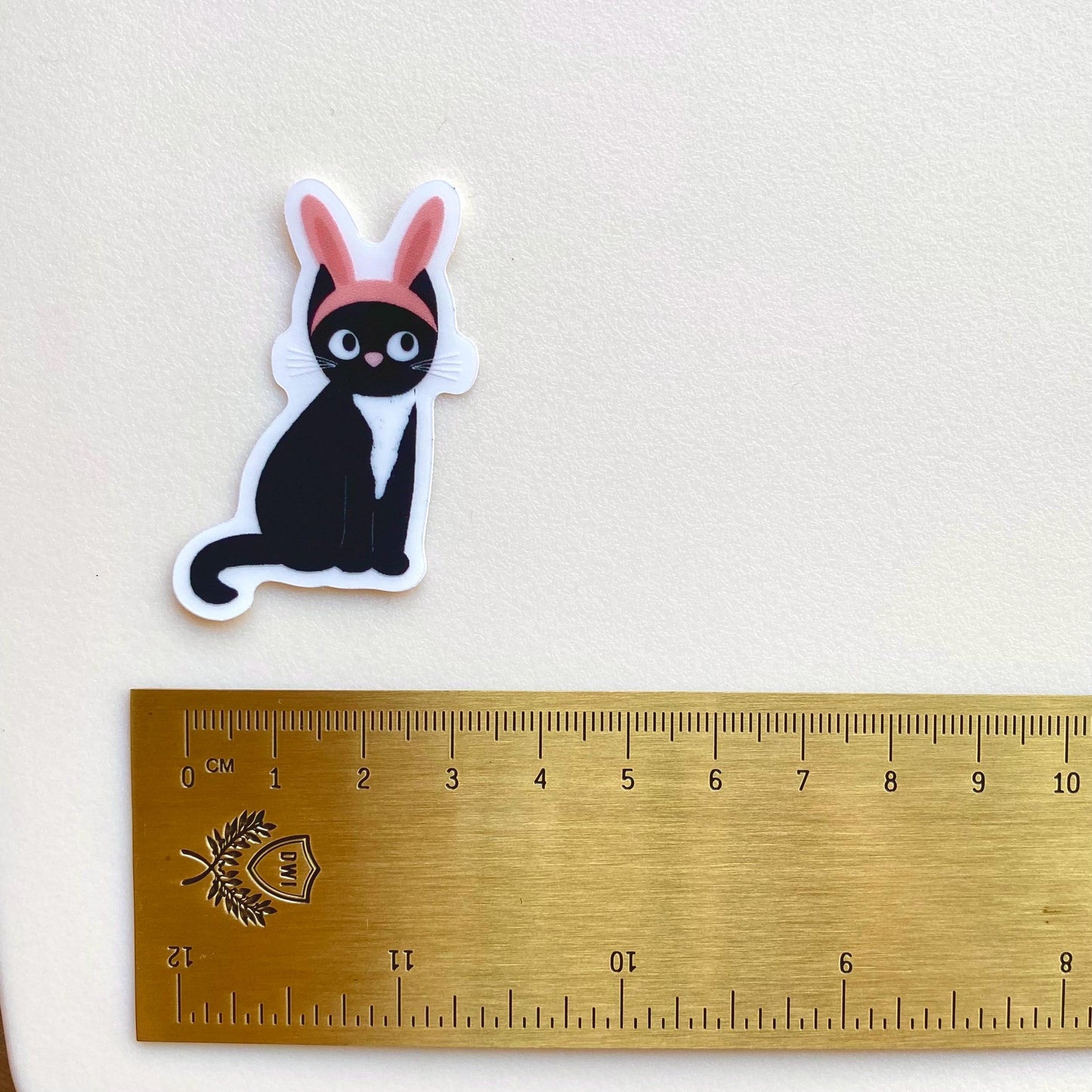 Sticker of a black cat with pink bunny ears next to a ruler on a beige background