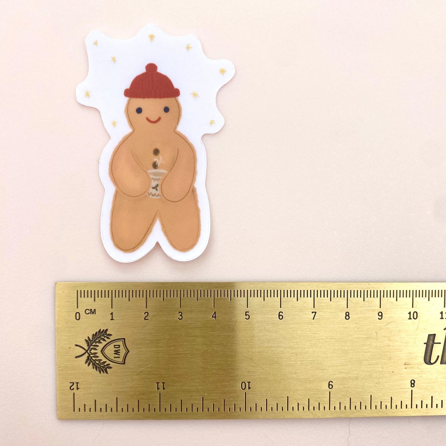 Sticker of a gingerbread man holding a festive hot chocolate mug next to a ruler for scale on a beige background