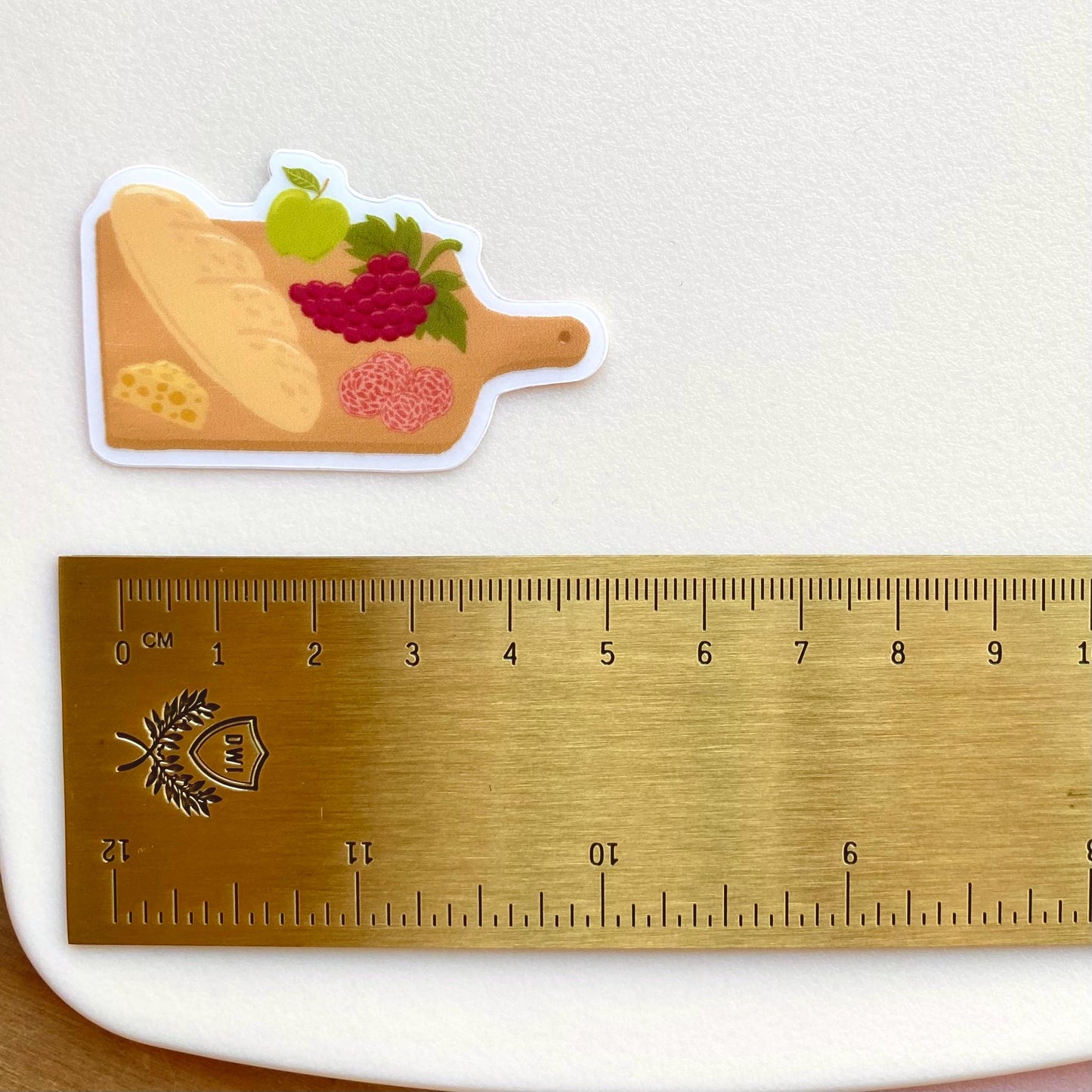 Charcuterie board sticker with a baguette, cheese, grapes, apple and salami, against a cream background with a gold ruler for scale.