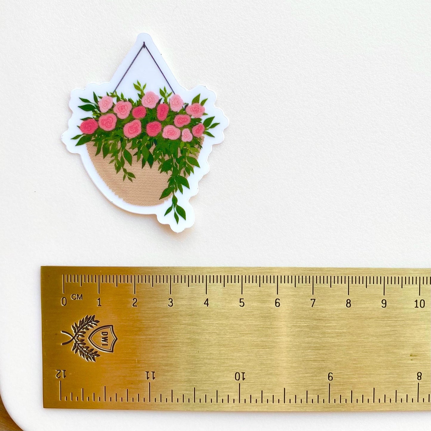A sticker with a hanging planter with pink flowers against a white background with a gold ruler for scale.