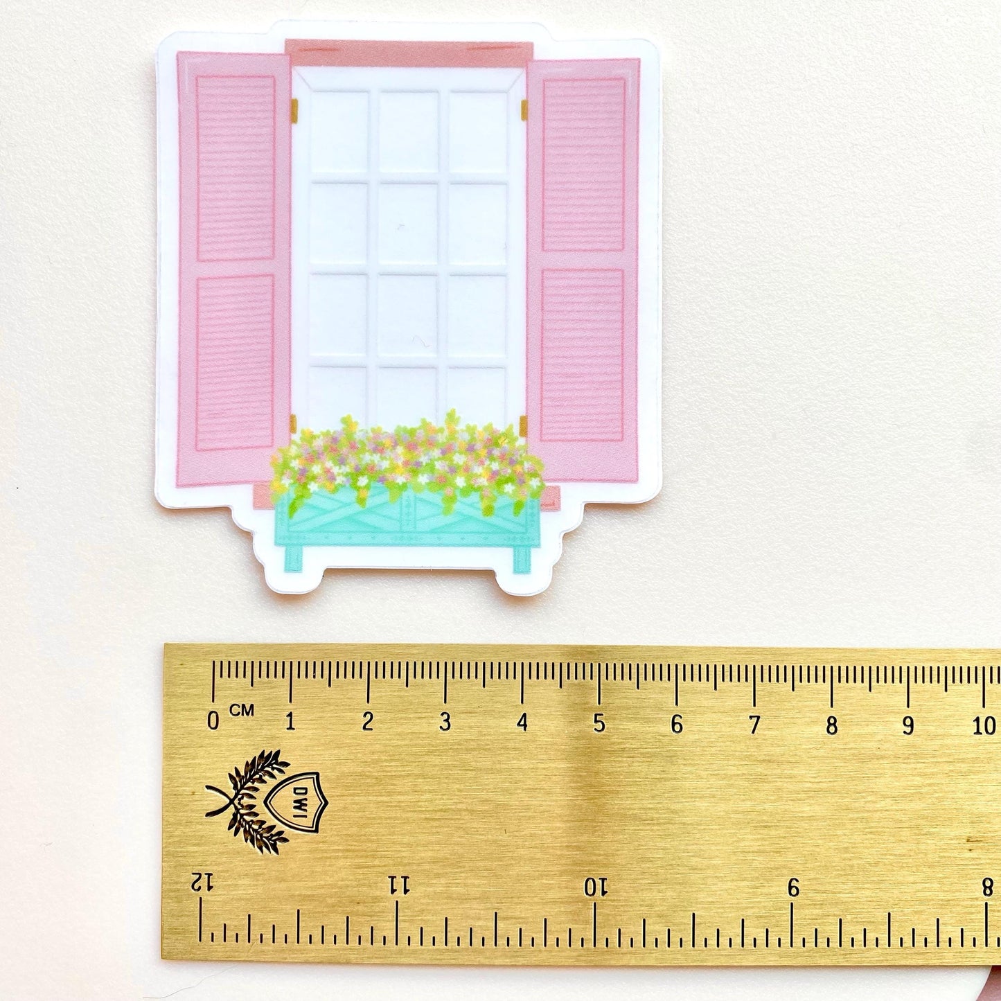 Transparent sticker of a window with pink shutters and a flower box on a white background with a gold ruler for scale.