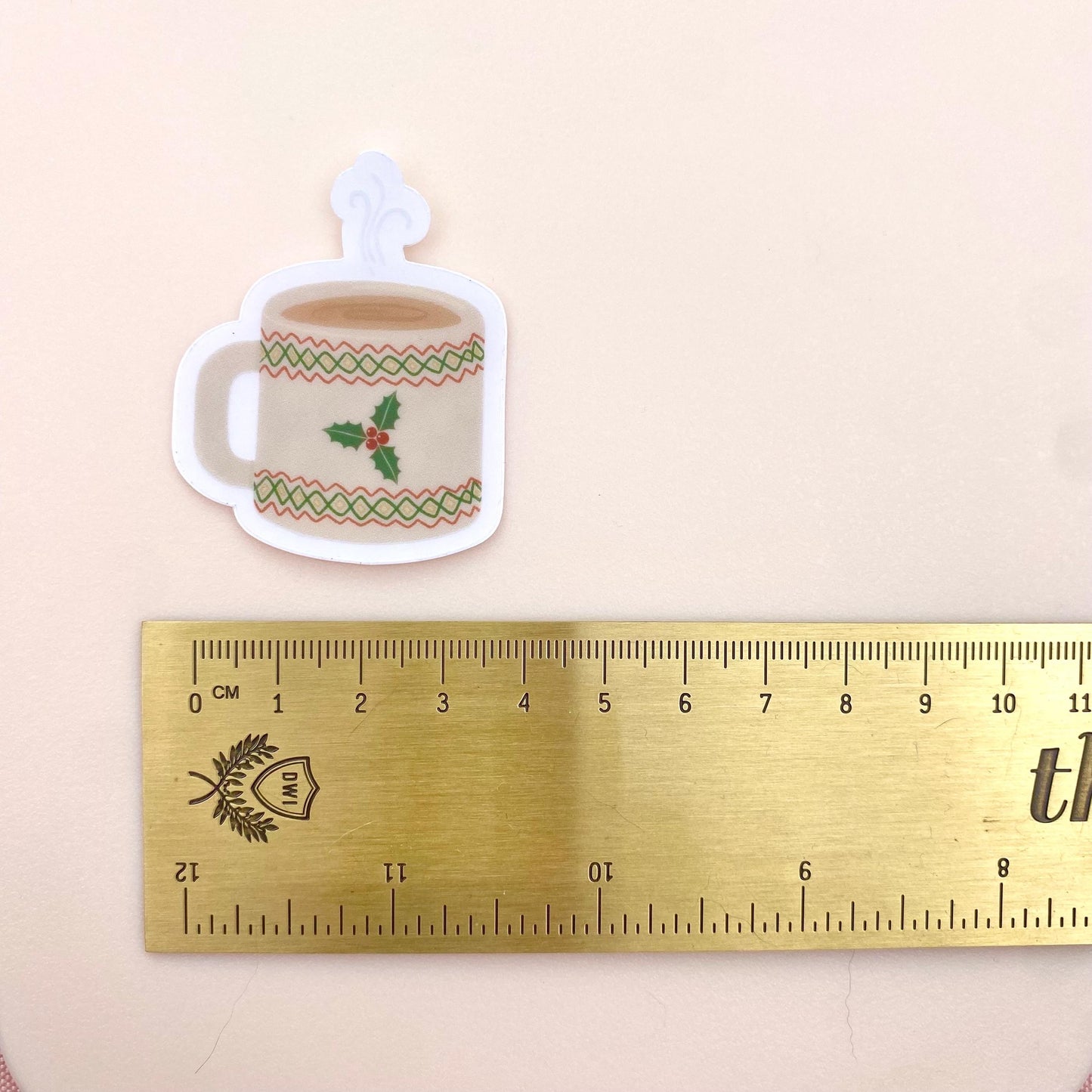 Sticker of a Christmas-themed mug with holly design next to a ruler for scale on a beige background