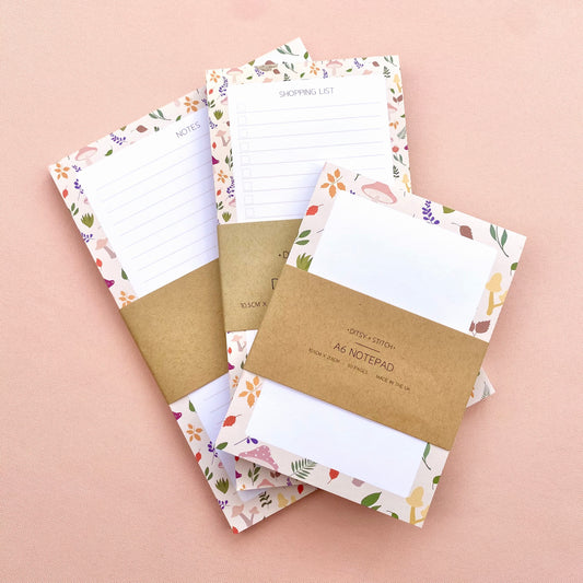 Three floral notepads with kraft paper bands, arranged on a soft pink background. The notepads have white lined writing pages and a delicate autumn botanical border.