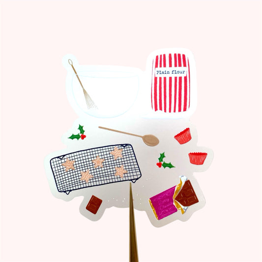A sticker with an illustration of christmas baking set with a red and white striped flour bag, wooden spoon, mixing bowl and whisk, chocolate and star shaped cookies, held up against a pink background.