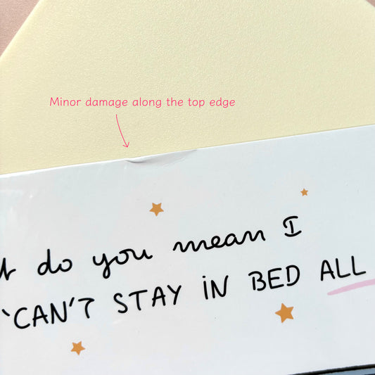 A close-up of a white illustration print with handwritten black text reading 'What do you mean I CAN'T STAY IN BED ALL DAY?' and small gold star accents. Pink annotation text says 'Minor damage along the top edge' with an arrow pointing to a crease.