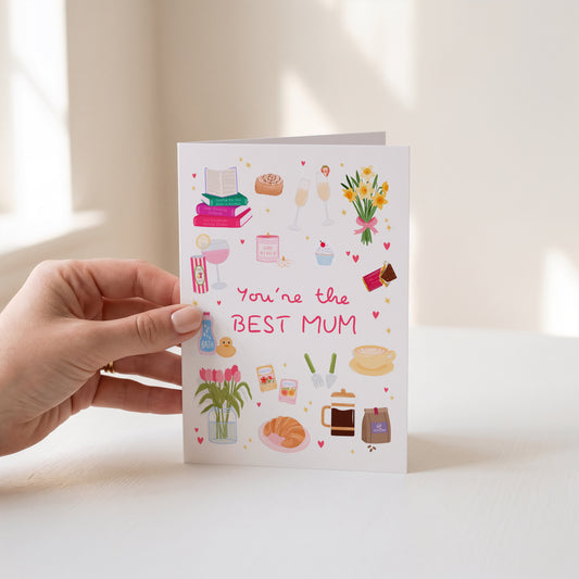 A hand holds a white greeting card that says 'You're the BEST MUM' in pink lettering, decorated with colourful illustrations of books, flowers, coffee, croissants, and small heart accents, against a softly lit cream background.