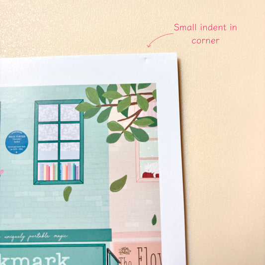 A close-up of a print showing part of an illustrated bookshop scene with green leaves, a window, and pastel colours, placed on a beige textured background. Pink text at the top reads 'Small indent in corner'.