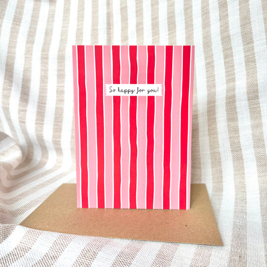 Red and pink striped card with 'So happy for you!' text on a brown kraft envelope against a striped fabric background.