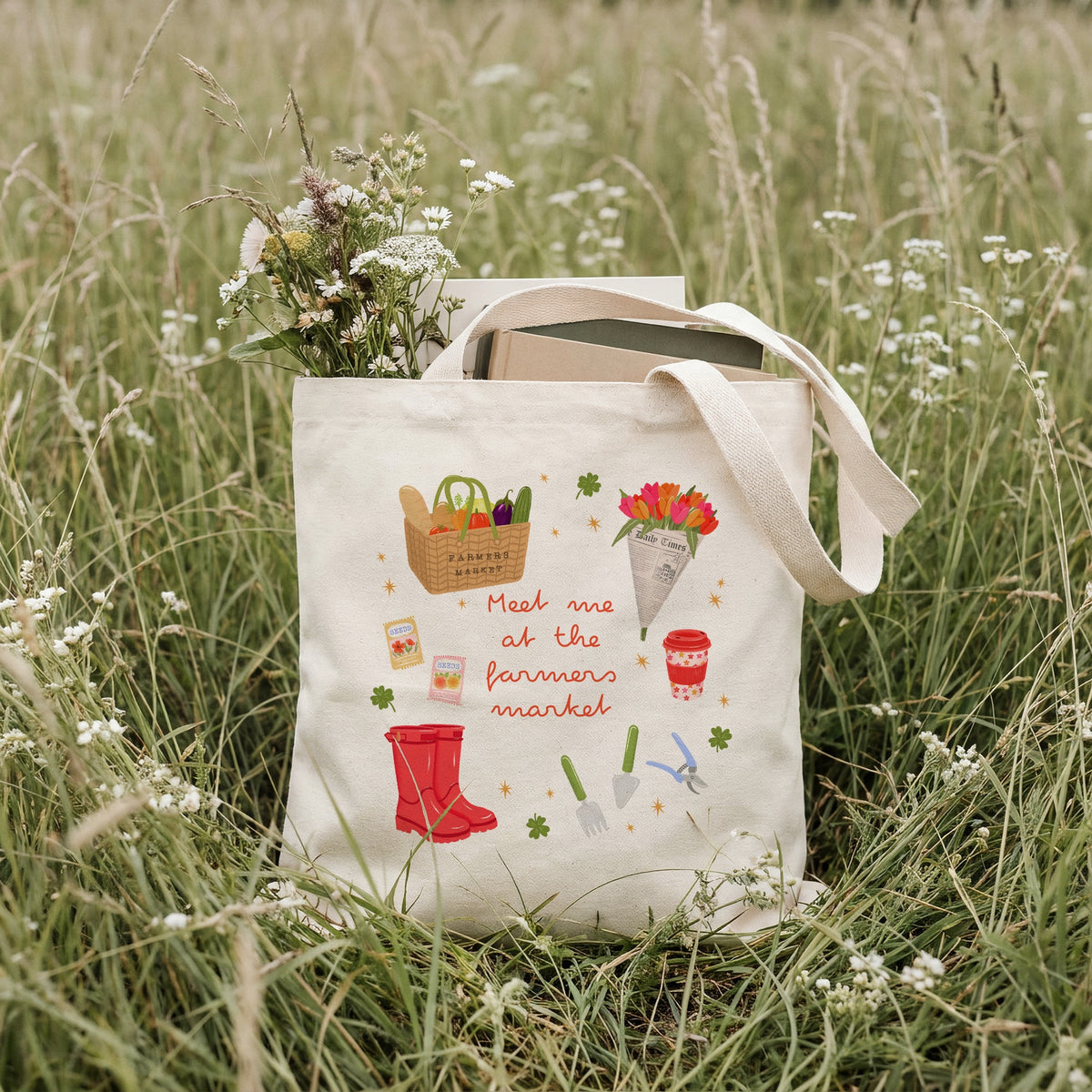 A cream canvas tote bag with a colourful 'Meet me at the farmers market' illustration, sitting in a grassy field with wildflowers. The bag contains books and a small bouquet of flowers.
