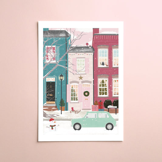 A vertical art print featuring a whimsical winter street scene with pastel-coloured buildings, a mint green car, and a snowman, displayed against a soft pink wall.