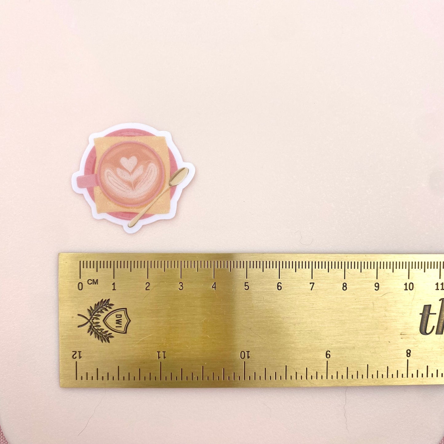 A transparent sticker with a detailed illustration of a latte from above with foamy heart shaped latte art, in a pink mug with a yellow napkin and gold teaspoon. Shown with a ruler for size reference.