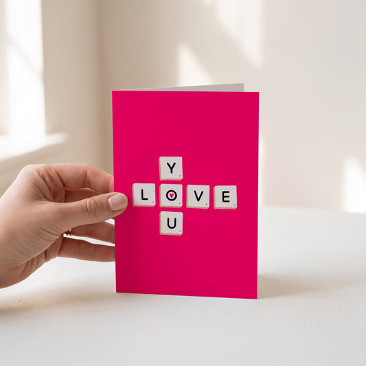 A bright pink greeting card held by a hand, featuring the word 'LOVE' spelled out with white letter tiles on the front, set against a softly lit neutral background.