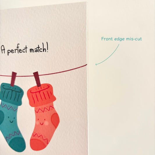A greeting card or print showing two cute sock illustrations hanging on a line, with the text 'A perfect match!' on the left and a blank off-white area on the right labeled 'Front edge mis-cut' with a teal arrow.