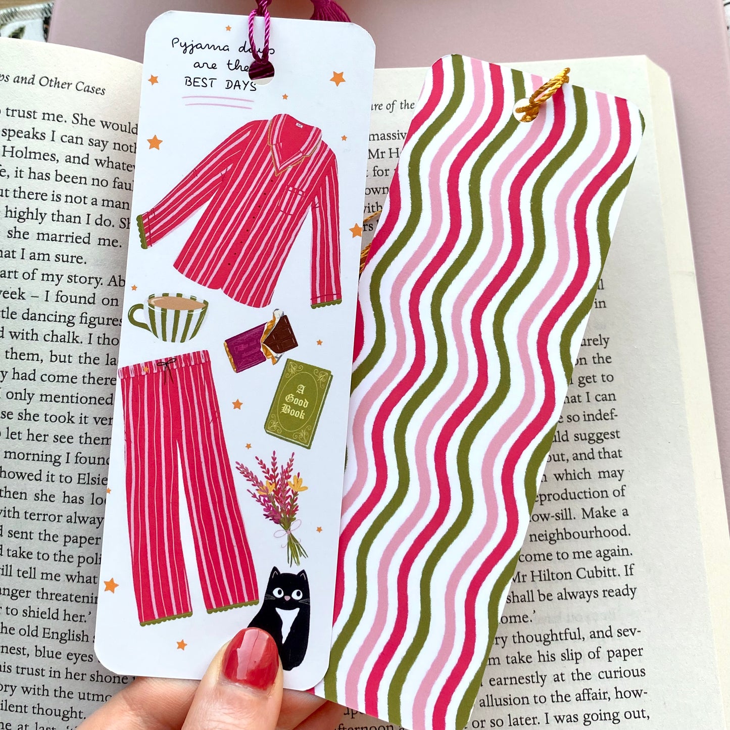 Two identical colourful bookmarks with one showing the front illustration of pink striped pyjamas, black cat, and book and the other showing the back wavy pattern. Shown on a book page.