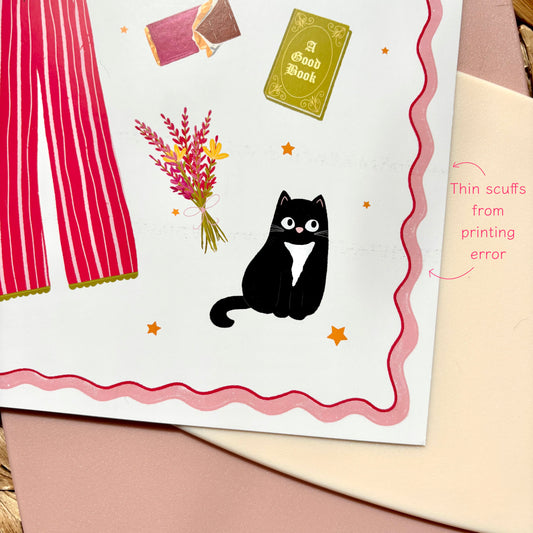 A close-up of a printed art print showing a whimsical illustration: a black cat with a white chest, pink curtains, flowers, stars, and a small book. A pink handwritten note reads 'Thin scuffs from printing error.