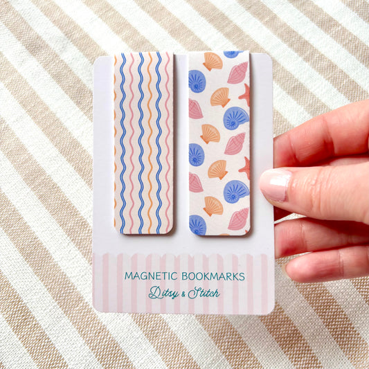 Two magnetic bookmarks with shell pattern design held by a hand on a striped background