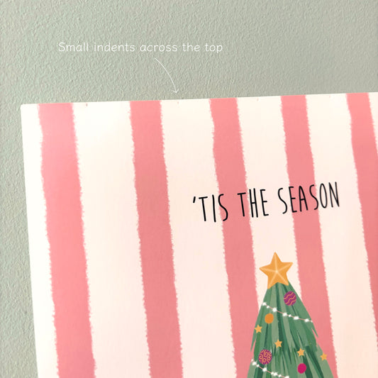 A close-up of an art print with pink and cream vertical stripes, the text “'TIS THE SEASON,” and part of a green Christmas tree illustration, shown against a pale green wall. A small handwritten note at the top says “Small indents across the top.