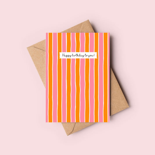 Greeting card with pink and orange stripes and 'Happy birthday to you!' text, placed on a beige envelope against a light pink background.