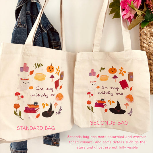 Two canvas tote bags displayed side by side against a white wall, with a denim jacket hanging on the left and flowers in a basket on the upper right. The left bag is labelled 'STANDARD BAG' and the right bag 'SECONDS BAG'; both are off-white with a whimsical witch-themed illustration including pumpkins, books, mushrooms, and a black hat, plus pink text reading 'In my witchy era'.