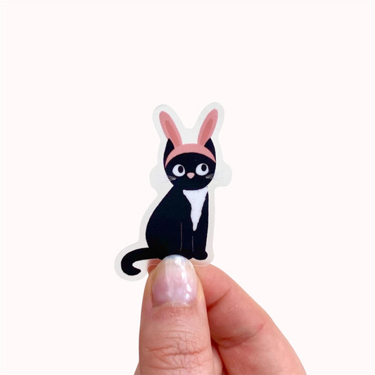 Sticker of a black cat with pink bunny ears held by a hand on a white background