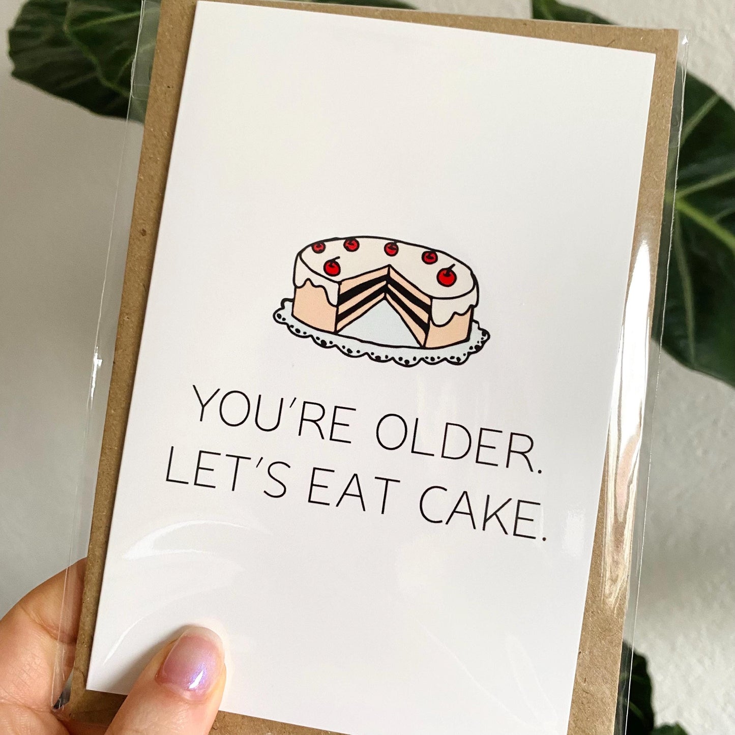 Greeting card with a cake illustration and text 'You're older. Let's eat cake.' held by a hand.