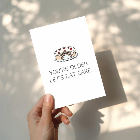 Hand holding a card with a cake illustration and text 'You're older. Let's eat cake.'
