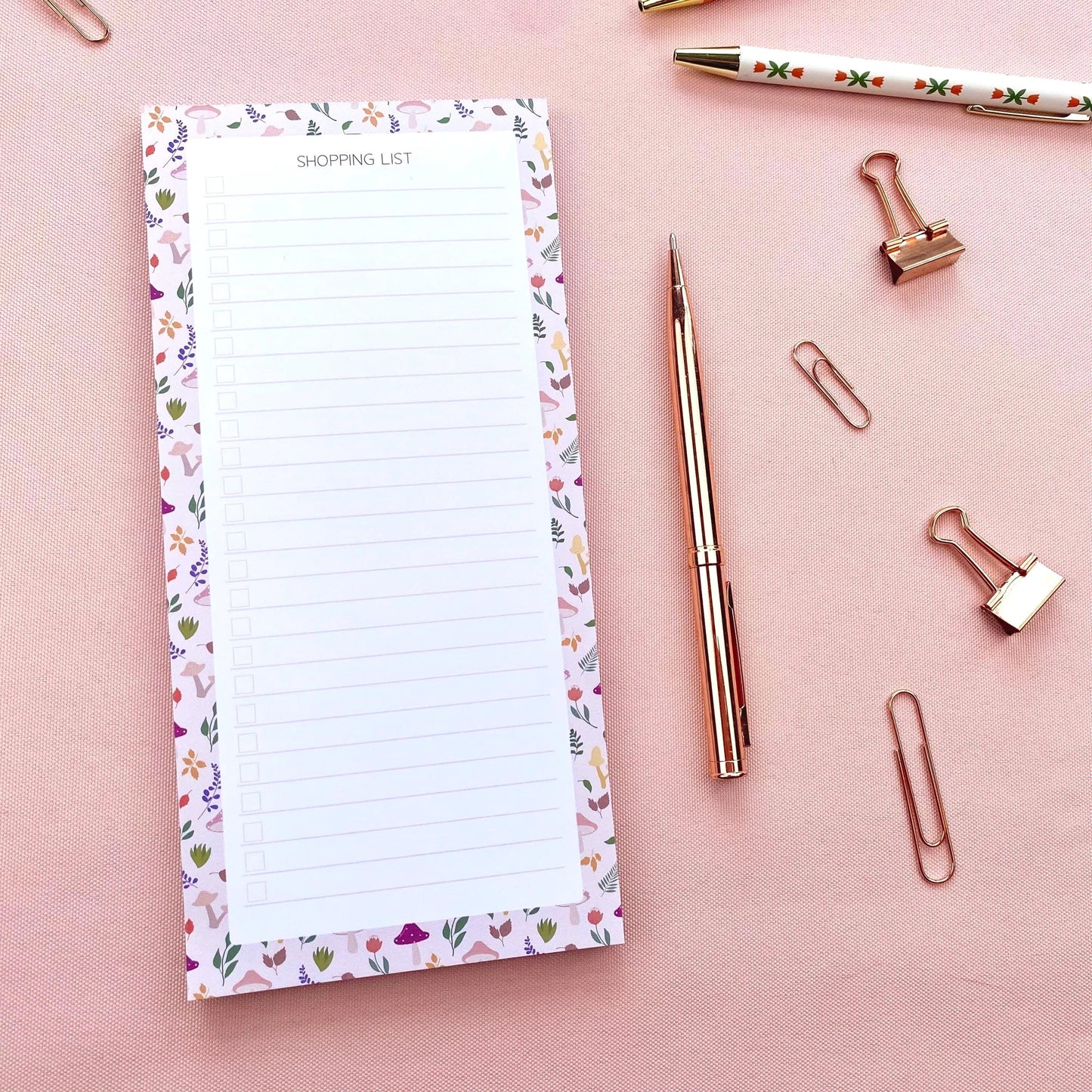 Autumn foliage-patterned shopping list pad with a pen and paper clips on a pink background