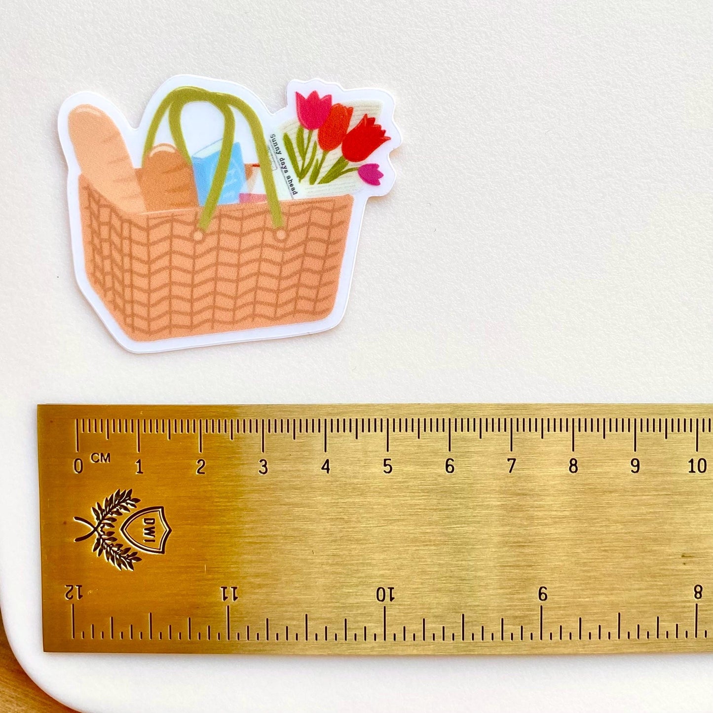 Sticker of a picnic basket with flowers next to a ruler for scale on a light background