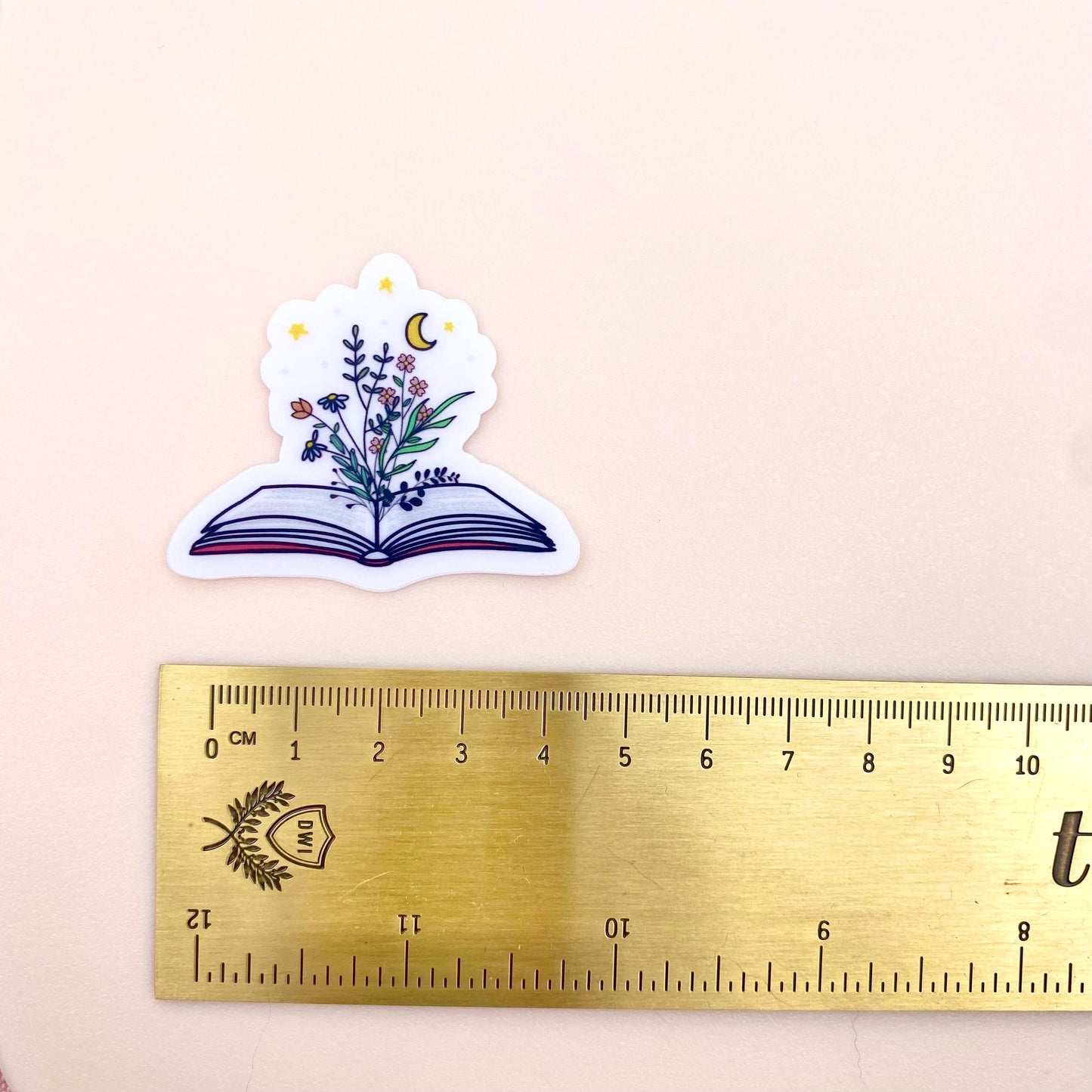 Decorative sticker of an open book with floral designs and a moon and stars on a white background with a gold ruler for scale