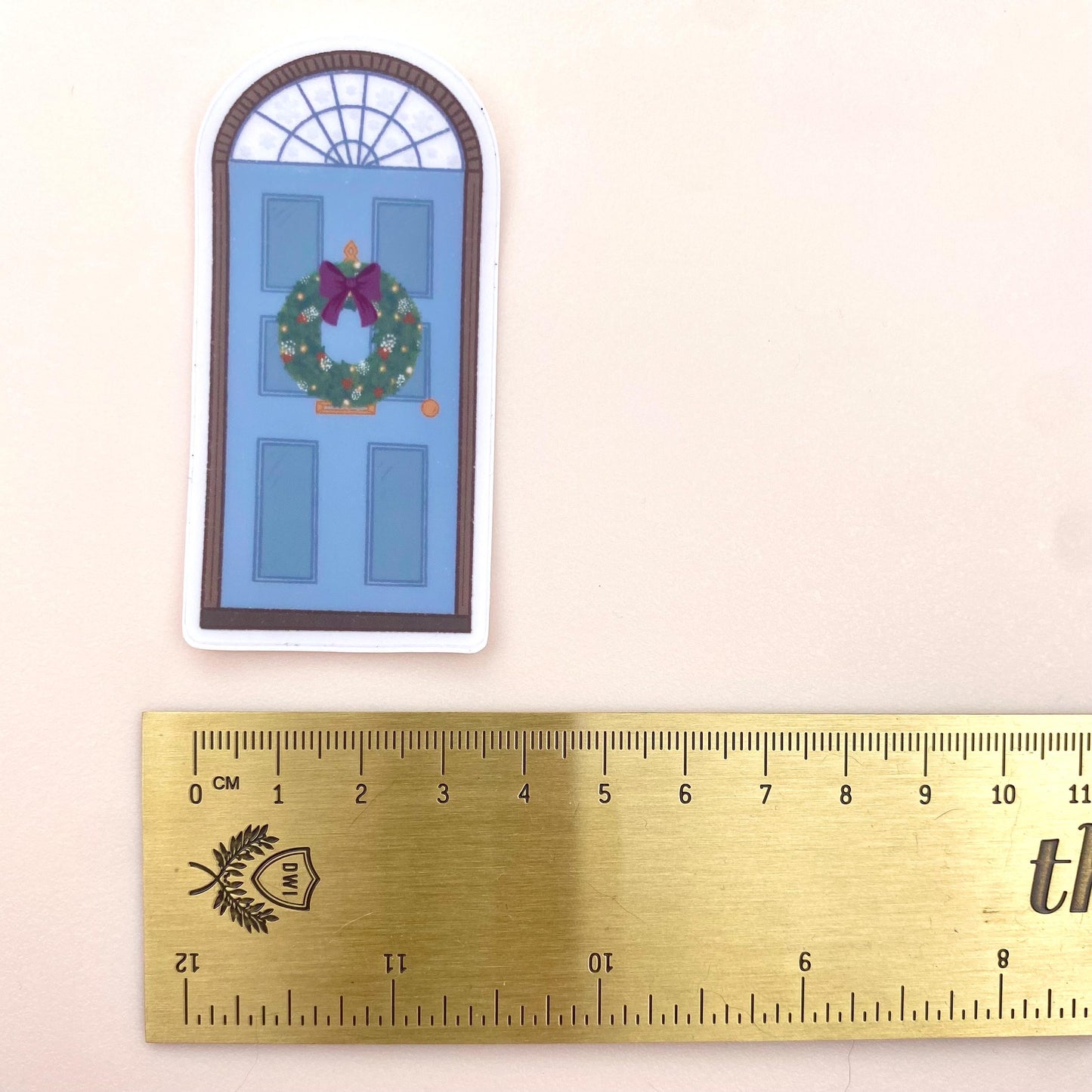 A sticker with a blue door decorated for Christmas against a cream background with a gold ruler for reference.