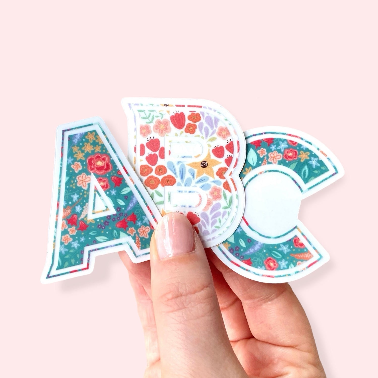 Hand holding 3 stickers with the letters 'A', 'B' and 'C' with a mix of floral patterns against a pink background