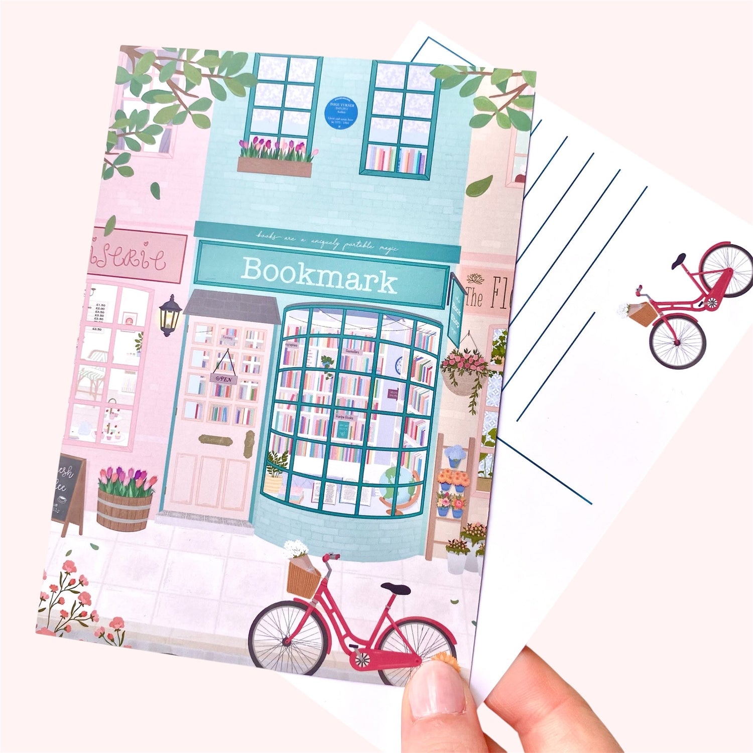 A hand holding a postcard with a bookshop illustration and a second postcard showing the back design with lines for writing and a pink bicycle illustration in the bottom right corner