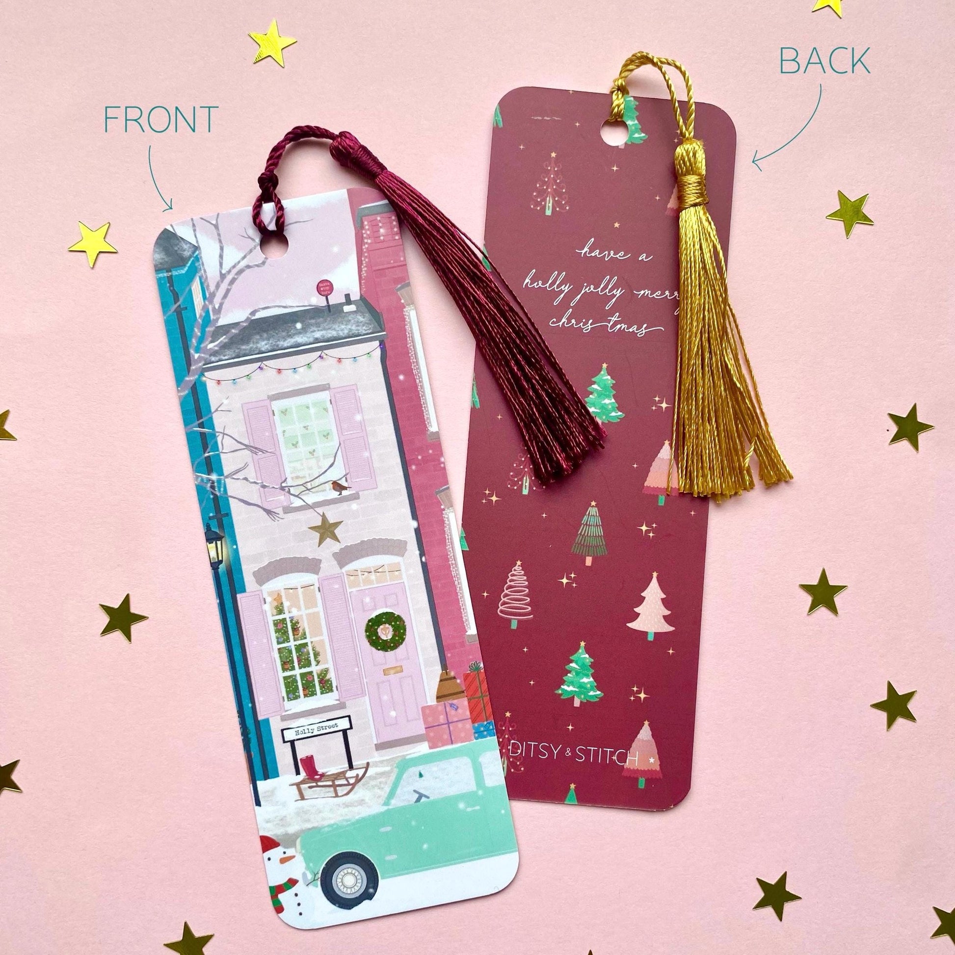 Decorative bookmark depicting a wintery street scene with pink houses with gold tassel on a pink background. Second bookmark showing design on the back with a dark pink Christmas trees pattern and holly jolly quote