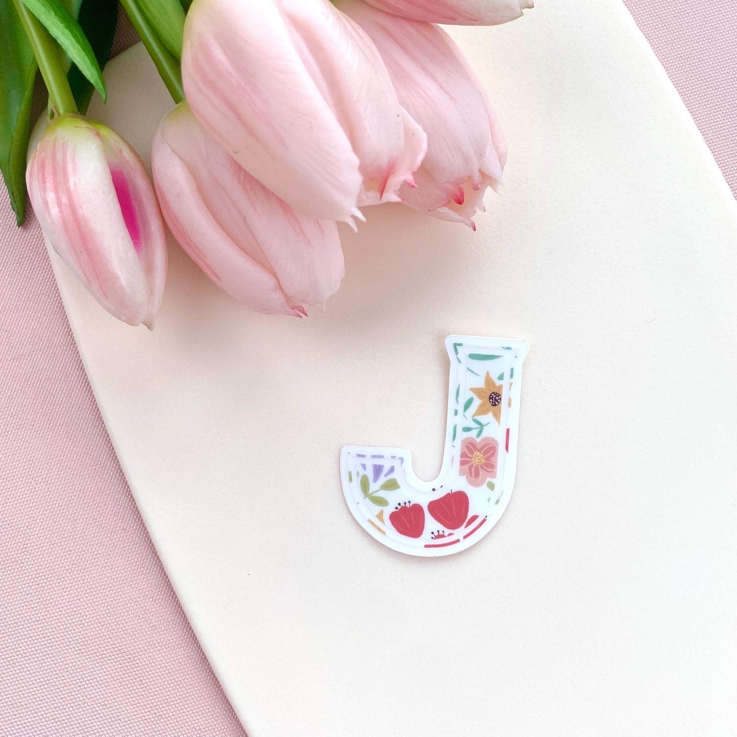 Floral-styled letter 'J' sticker on a white surface with pink tulips in the background.