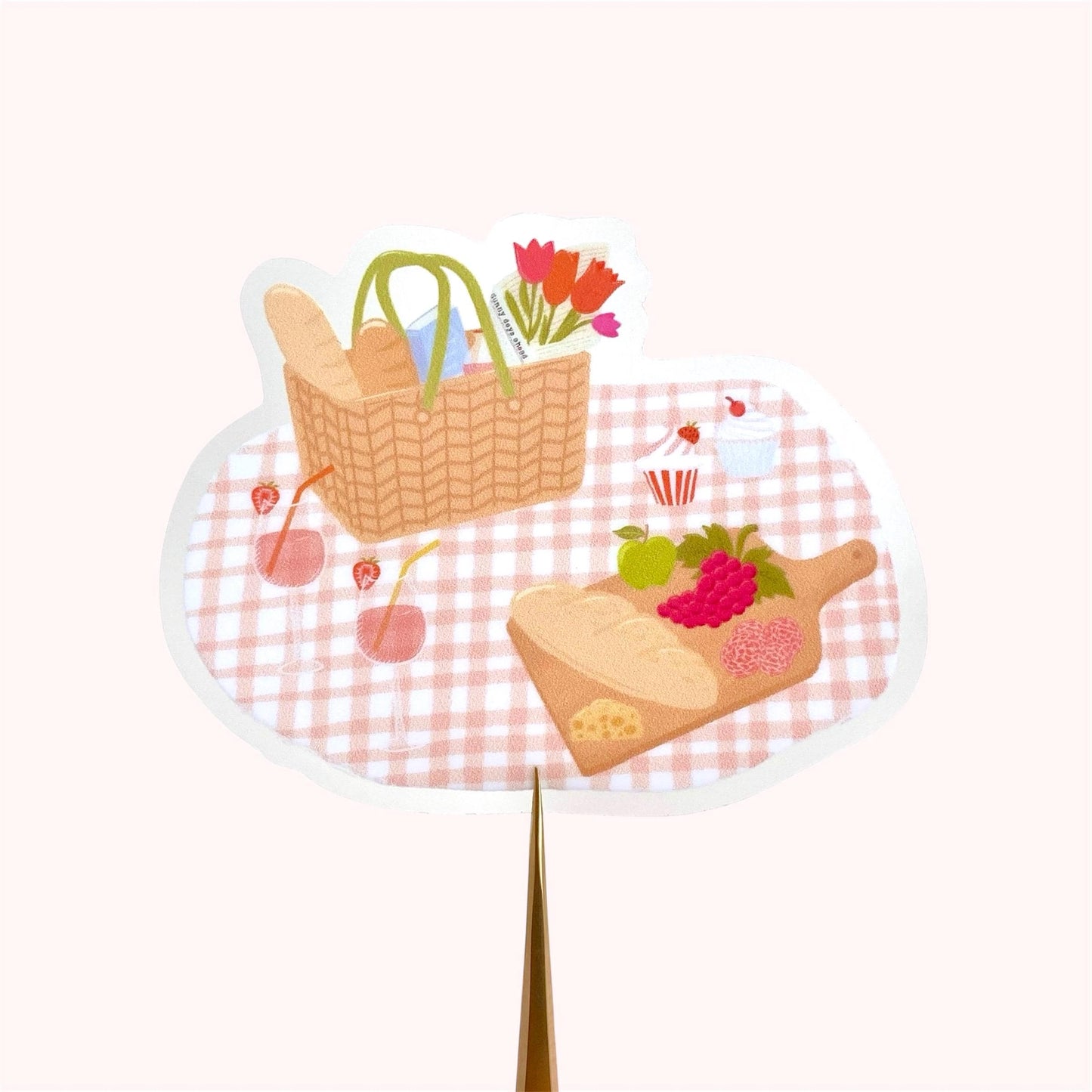 Sticker with an illustration of a picnic scene with a basket, bread, and flowers on a checkered tablecloth.