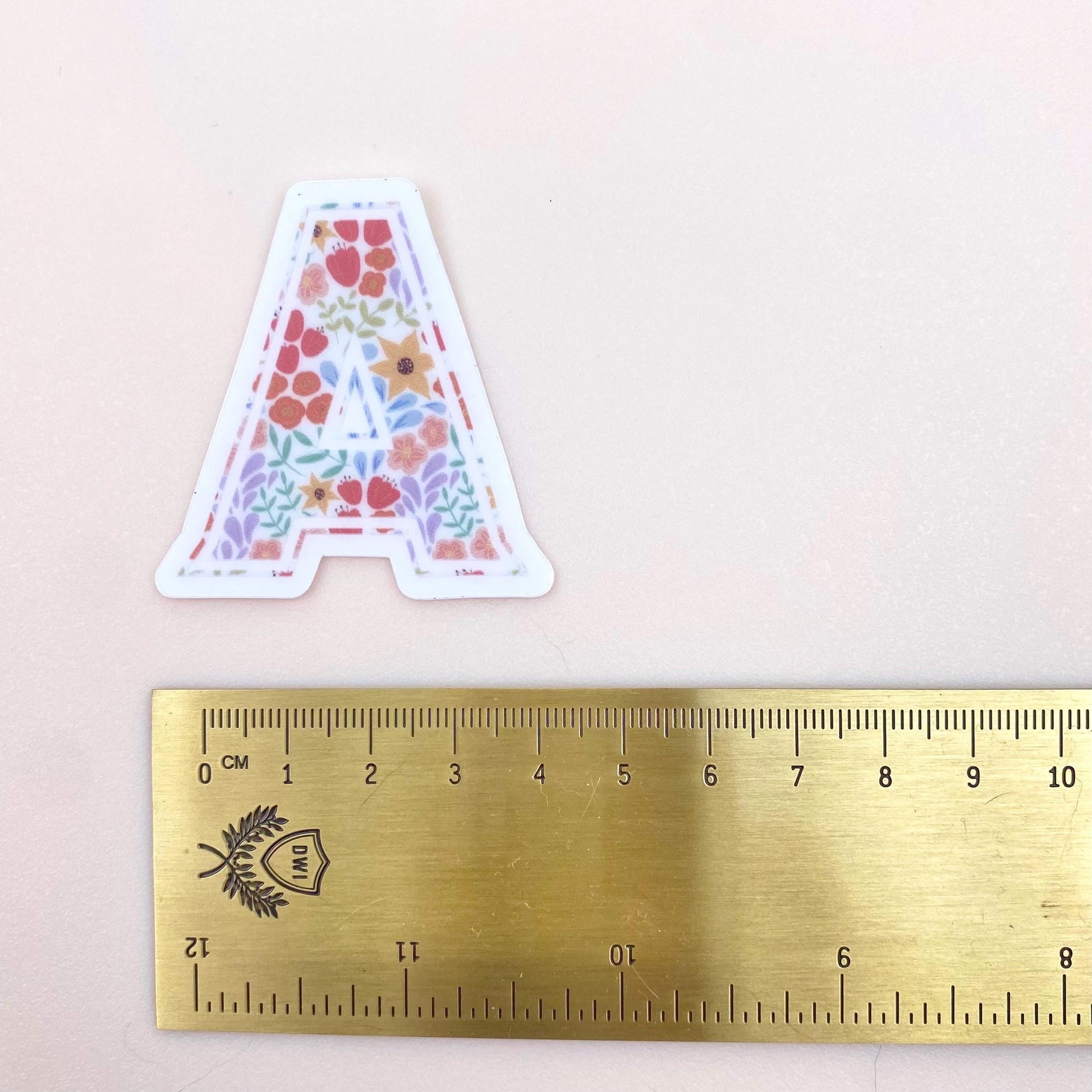 Floral letter 'A' sticker next to a ruler on a light pink background