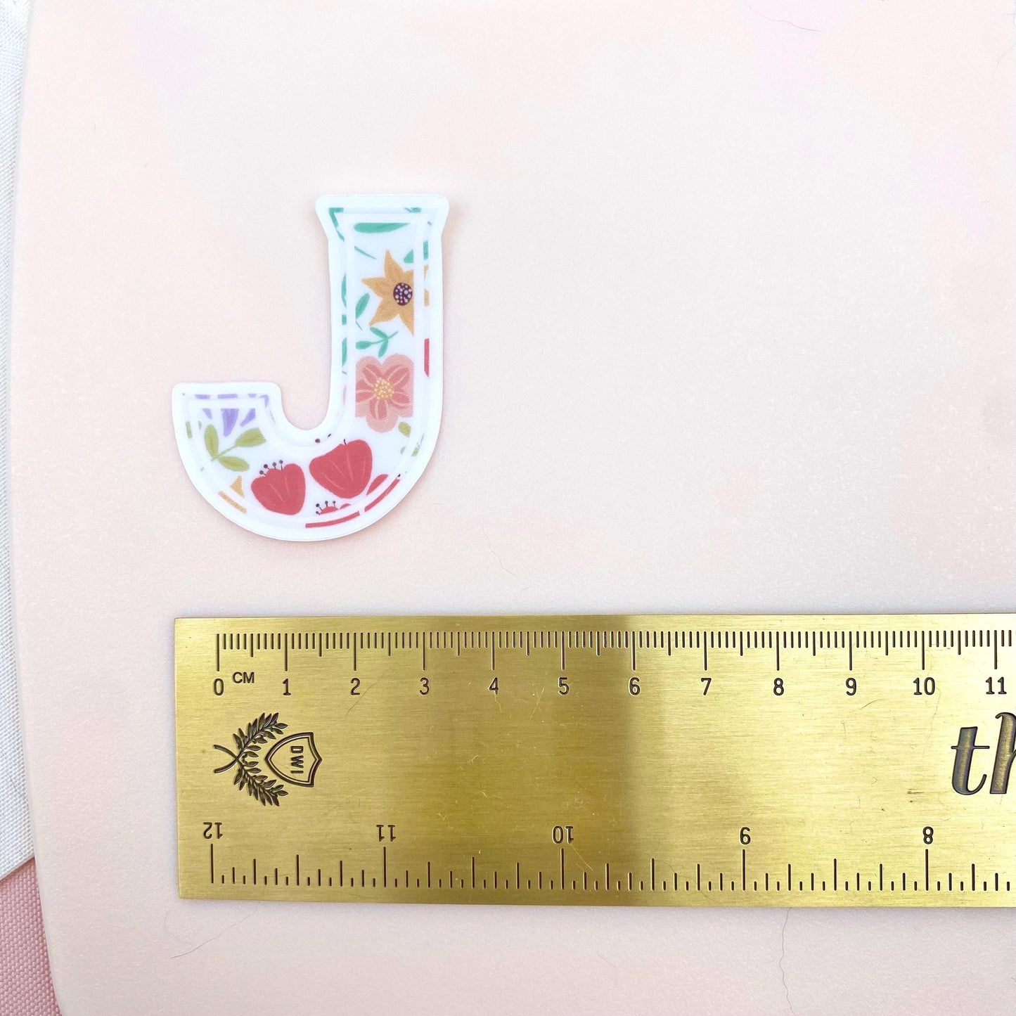 Floral letter 'J' sticker next to a ruler on a pink background