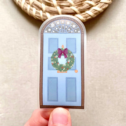 A sticker with a blue door decorated for Christmas held by hand against a neutral background.