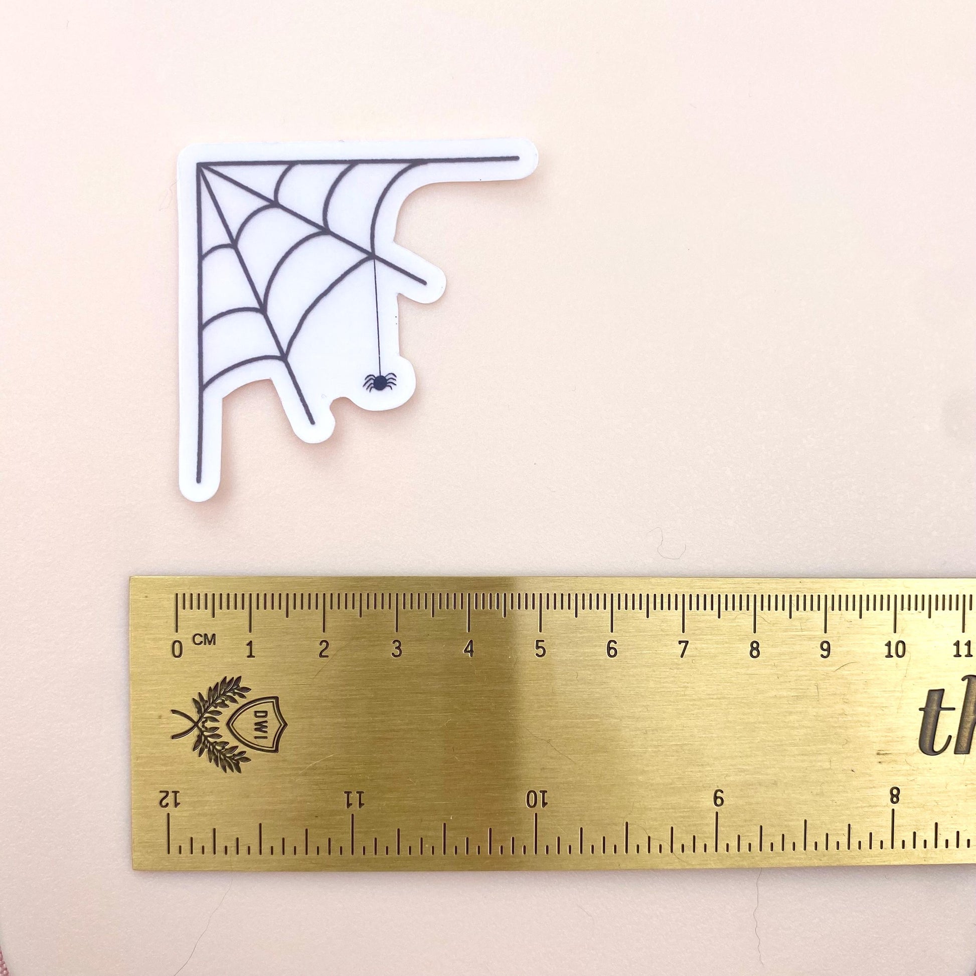 Transparent sticker with corner spider web design with a hanging spider on a white background with a gold ruler for scale.