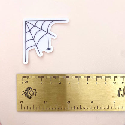 Transparent sticker with corner spider web design with a hanging spider on a white background with a gold ruler for scale.