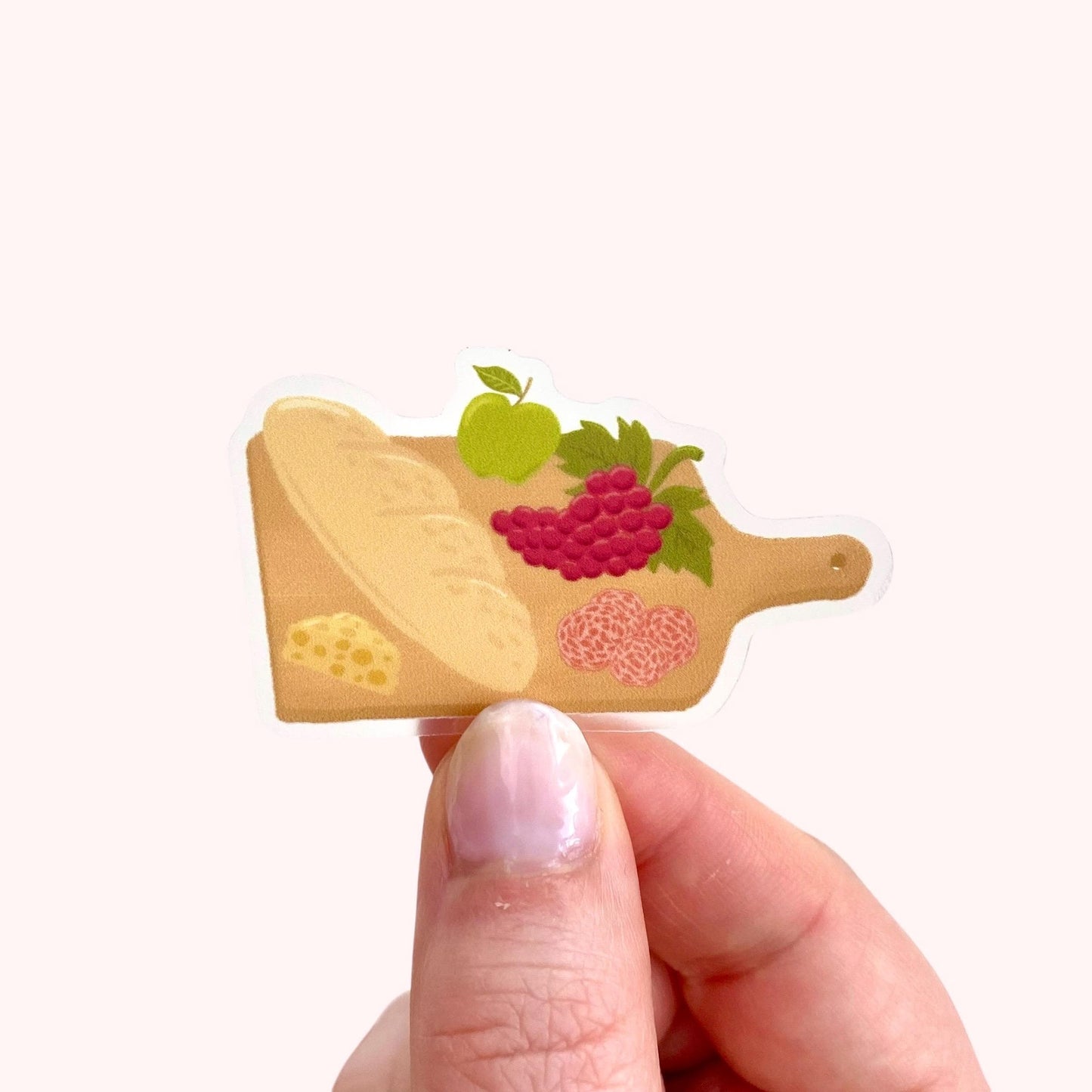 Charcuterie board sticker with a baguette, cheese, grapes, apple and salami, held by hand against a cream background