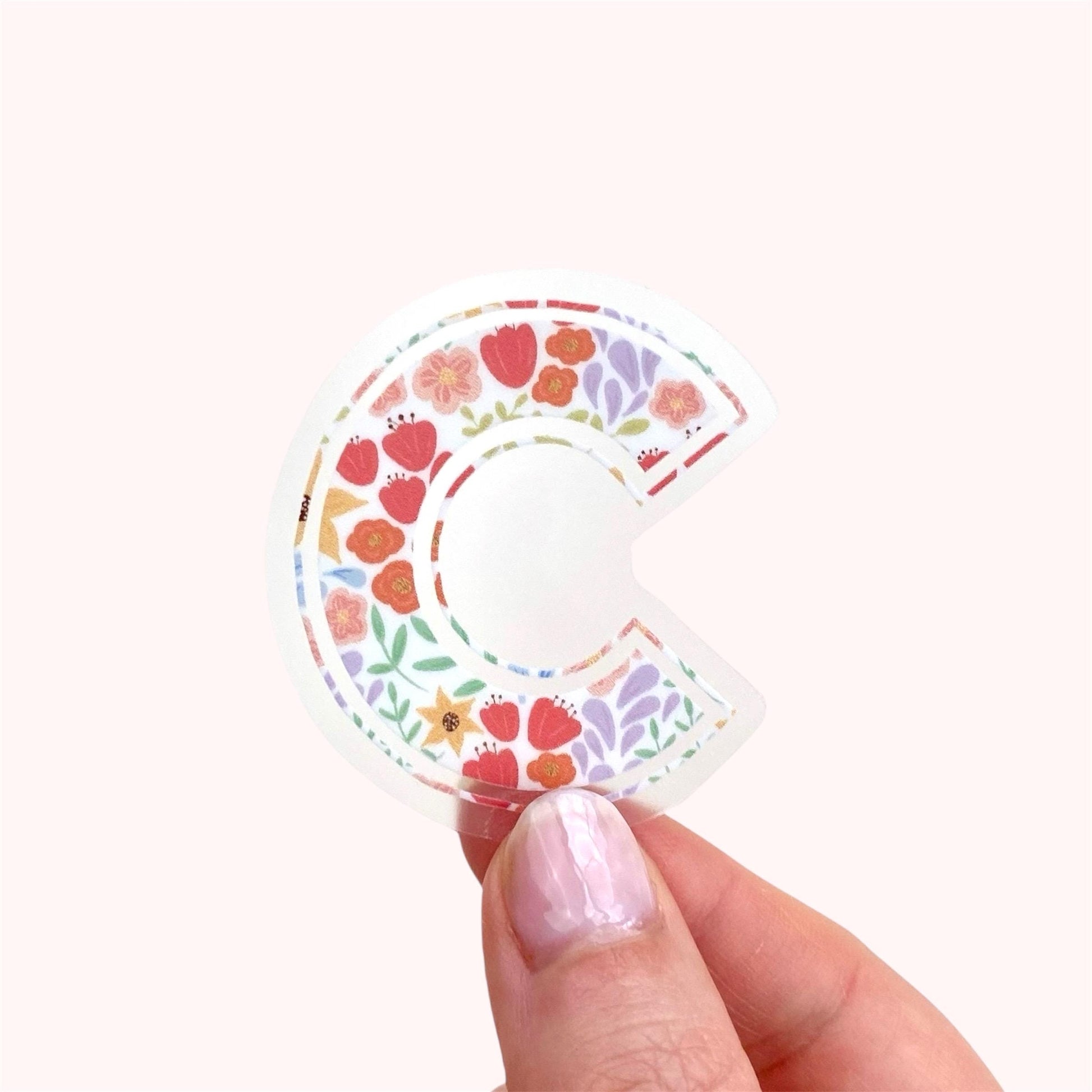 Decorative letter 'C' sticker with floral design held by a hand on a white background